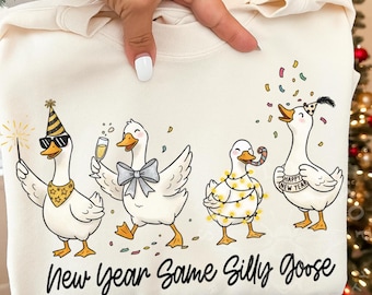 May include: Cream-colored sweatshirt featuring a festive goose design. Four cartoon geese are depicted celebrating with party hats, sparklers, champagne, and confetti. The text "New Year Same Silly Goose" is printed below the geese.