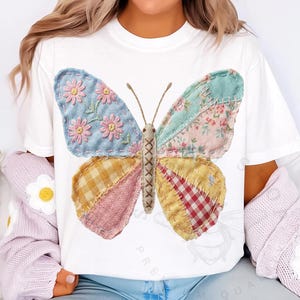 May include: White t-shirt featuring a large, colorful butterfly design. The butterfly has patchwork wings with floral and plaid patterns in shades of blue, pink, yellow, and red. The butterfly's body is tan, and the antennae are brown.