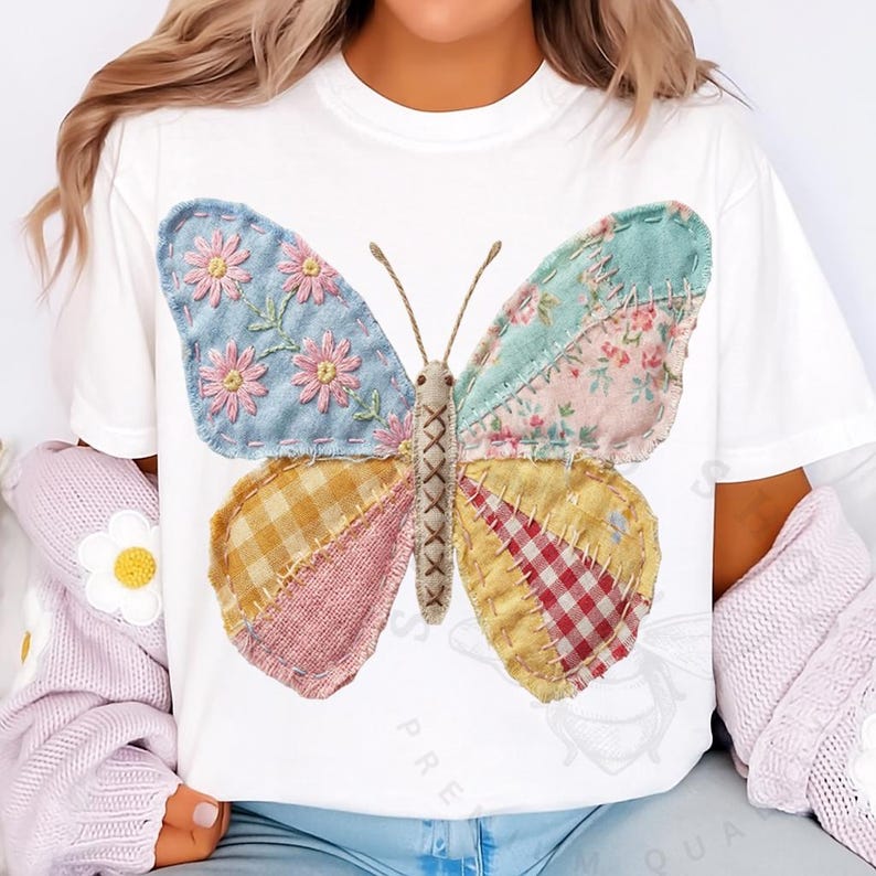 May include: White t-shirt featuring a large, colorful butterfly design. The butterfly has patchwork wings with floral and plaid patterns in shades of blue, pink, yellow, and red. The butterfly's body is tan, and the antennae are brown.