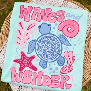 Pu&ograve; includere: T-shirt azzurra con le parole "WAVES and WONDER" in lettere rosa a bolle. La maglietta presenta una tartaruga blu, una stella marina rosa e altre grafiche a tema marino.