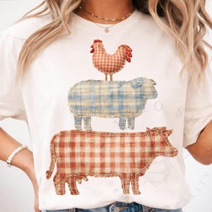 May include: White t-shirt featuring a stacked farm animal design. A red and white gingham cow is at the bottom, a blue and white gingham sheep in the middle, and a red and white gingham rooster at the top. The design is a patchwork style.
