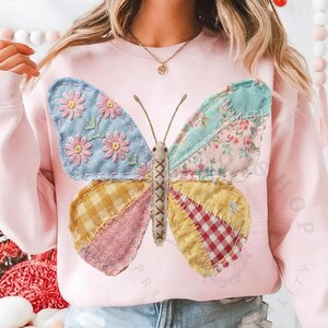 May include: A light pink sweatshirt featuring a patchwork butterfly design. The butterfly wings are made of various patterned fabrics in blue, yellow, pink, and floral prints. The butterfly's body is stitched with a cross-stitch pattern.