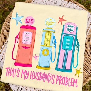 May include: A light yellow t-shirt featuring a graphic of three vintage gas pumps in pink, blue, and teal, with star accents. The text "THAT'S MY HUSBAND'S PROBLEM" is written in pink script across the bottom.