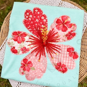 May include: A light blue t-shirt featuring a large, appliqued hibiscus flower. The flower petals are made from various patterned fabrics in red and pink, with a red embroidered center and yellow stamens. The shirt is displayed on a wicker basket.