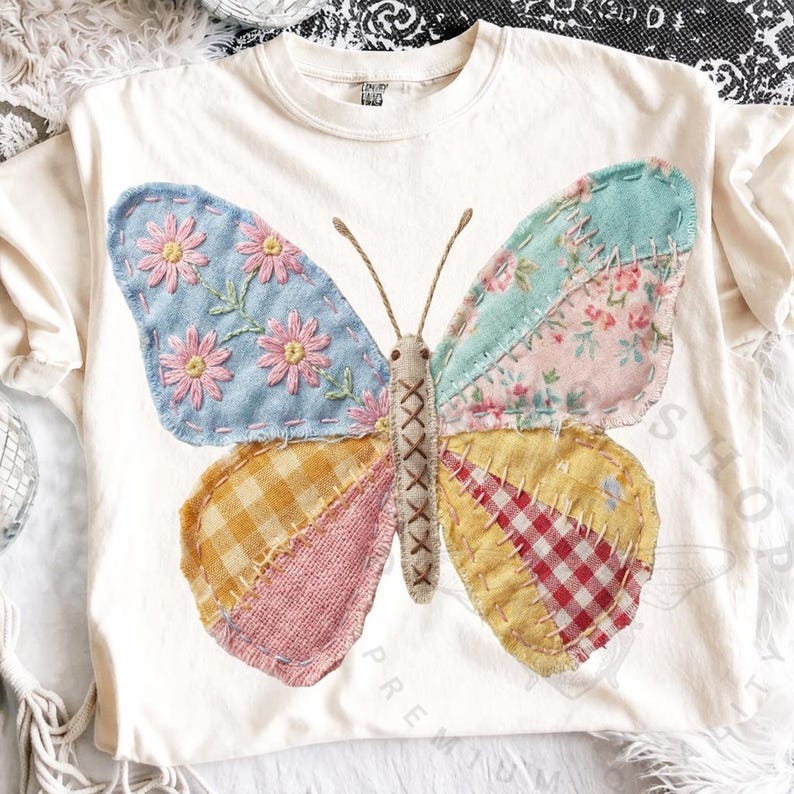 May include: Cream-colored t-shirt featuring a large, patchwork butterfly design. The butterfly has wings in various patterns, including floral, gingham, and plaid, with embroidered details. The butterfly's body is stitched with a cross-stitch pattern.