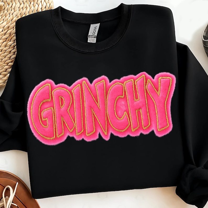 May include: Black sweatshirt with the word "GRINCHY" in large, pink, fuzzy letters outlined in gold. The sweatshirt has a crew neck and long sleeves. The word "GRINCHY" is the focal point of the design.
