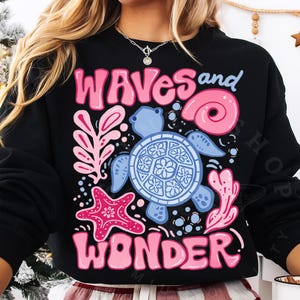 Pu&ograve; includere: Felpa nera con un design grafico colorato. Il design presenta le parole "WAVES and WONDER" in rosa, insieme a una tartaruga marina blu, una stella marina rosa e altri elementi a tema marino.