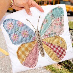 May include: A decorative butterfly applique on a white background. The butterfly features patchwork wings in various patterns and colors, including floral, gingham, and solid fabrics. The butterfly's body is a tan color with a stitched design, and the wings are adorned with embroidered details.