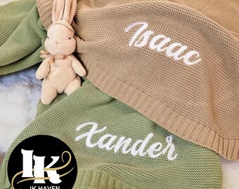 May include: Two personalized baby blankets, one tan and one green, with white embroidered names. A plush bunny toy with a bow sits on the blankets. The blankets are knitted and have a ribbed border. The logo for IK HAVEN is in the lower left corner.