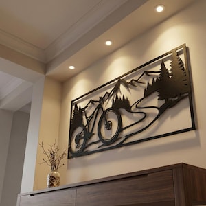 Peut inclure: Décoration murale en métal noir représentant un VTT sur un sentier, avec des montagnes et des arbres en arrière-plan. L'œuvre d'art rectangulaire est fixée sur un mur beige au-dessus d'un meuble en bois foncé.