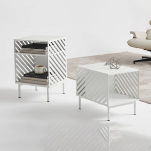 Peut inclure: Deux tables d'appoint en métal blanc avec un design à lattes diagonales. La table la plus haute possède deux étagères contenant des livres et une tasse en céramique. La table basse a une surface ouverte avec une sculpture géométrique en fil.