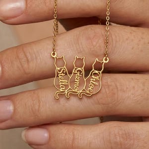 May include: A gold-colored necklace featuring three cat silhouettes. Each cat has a name engraved: Stella, Gizmo, and Chloe. The necklace is held by a person.