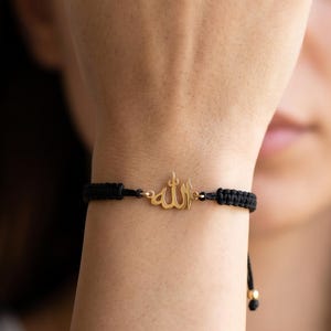 Puede incluir: Una pulsera de cordón negro con un dije de metal dorado en el centro. El dije muestra la caligrafía árabe para Allah. La pulsera está construida con un diseño de cordón trenzado e incluye pequeñas cuentas doradas en los extremos de los lazos.