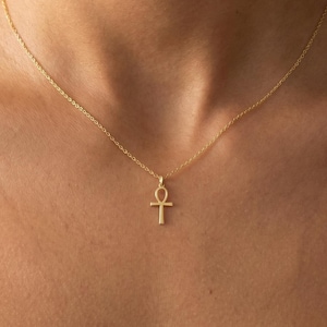 Peut inclure: Un délicat collier doré avec un pendentif Ankh. Le pendentif est une croix stylisée avec une boucle en haut. Le collier est porté autour du cou, et la chaîne est fine et subtile.