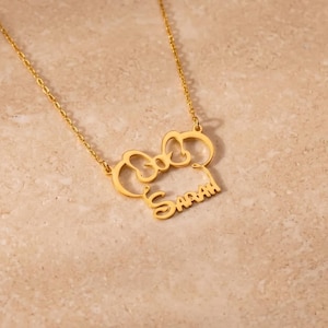 May include: A gold-colored necklace with a bow-shaped pendant and the name "Sarah" in a script font. The necklace is displayed on a light-colored, textured surface. The chain is delicate and the pendant is detailed.