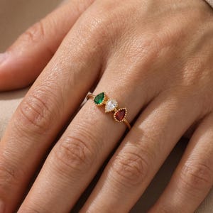 Peut inclure: Une bague en or ornée de trois pierres précieuses en forme de poire disposées en ligne. Les pierres comprennent une gemme verte à gauche, une gemme blanche transparente au centre et une gemme rouge à droite, serties dans un chaton doré perlé.