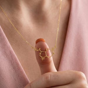 Può includere: Delicata collana color oro con un ciondolo a forma di struttura molecolare con dettagli floreali. Il ciondolo è sospeso a una catena sottile. La collana è mostrata su uno sfondo rosa.