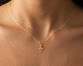 Pode incluir: Um delicado colar dourado com um pingente Ankh. O pingente é uma pequena cruz estilizada com uma alça na parte superior. O colar é usado ao redor do pescoço, e a corrente é fina e elegante.