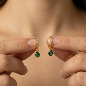 May include: A pair of gold-colored hoop earrings with small clear stones and teardrop-shaped, dark green gemstones. The earrings are held up against a blurred background.