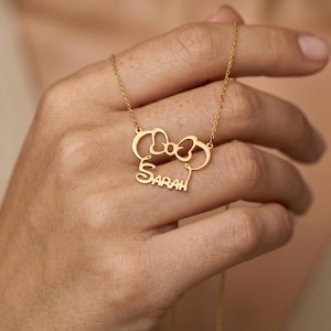 May include: A gold-colored necklace featuring a bow and mouse ears design with the name "Sarah" in a script font. The pendant hangs from a delicate chain. The necklace is held by a person.