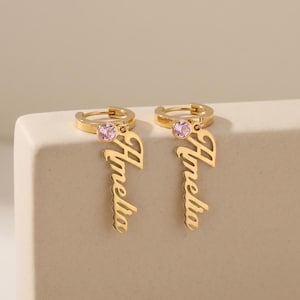 Könnte beinhalten: Ein Paar goldfarbene Creolen mit einem hängenden Namensanhänger, der den Namen Amelia in Schreibschrift zeigt. Jeder Ohrring ist mit einem kleinen, runden, hellrosa Edelstein verziert, der in einer runden Fassung am Ohrring befestigt ist.