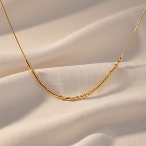 Könnte beinhalten: Eine zarte goldene Halskette mit einer Reihe kleiner Perlen und zylindrischen Stäben. Die Halskette wird auf einem weißen, strukturierten Stoff präsentiert. Die Kette ist dünn und das Design ist minimalistisch und elegant.