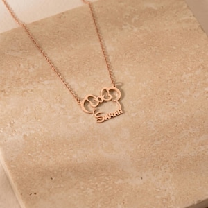 May include: Rose gold necklace featuring a bow-shaped pendant with the name "Sarah" in a delicate font. The necklace is displayed on a textured, neutral-toned surface, highlighting the jewelry's design and color.