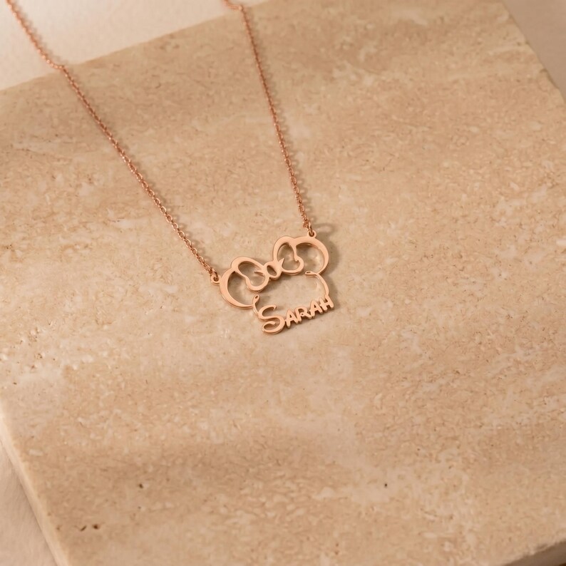May include: Rose gold necklace featuring a bow-shaped pendant with the name "Sarah" in a delicate font. The necklace is displayed on a textured, neutral-toned surface, highlighting the jewelry's design and color.