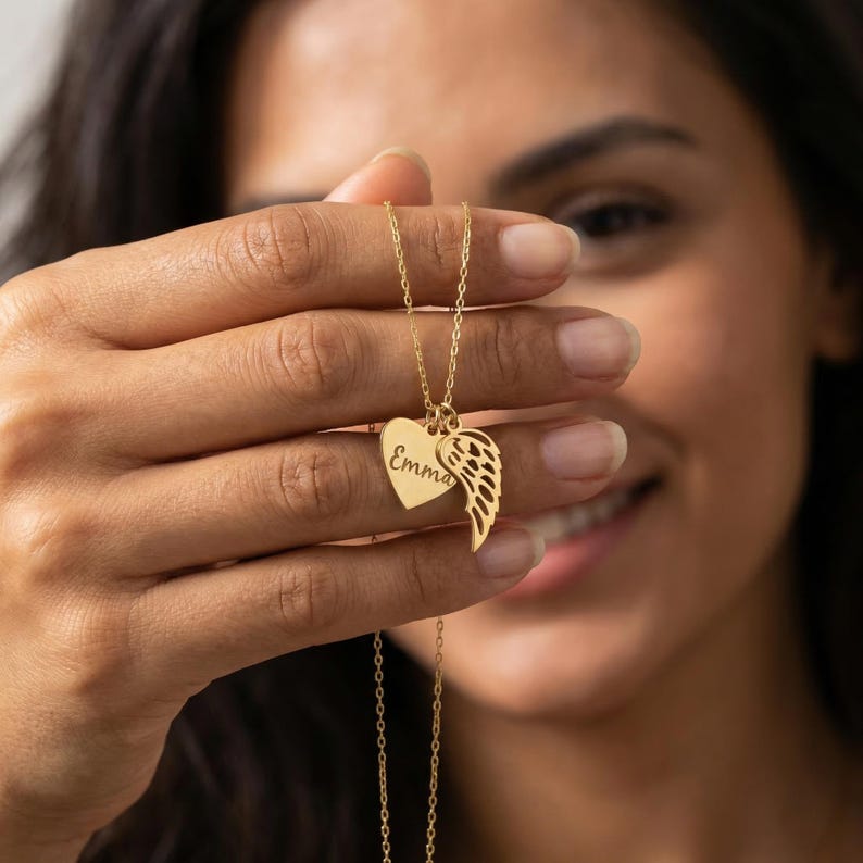May include: A gold-colored necklace with a heart-shaped pendant and a wing charm. The heart pendant has the name "Emma" engraved on it. The necklace is held up by a person.