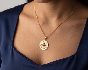 May include: Gold-colored compass pendant necklace. The pendant is round with a detailed compass rose design. The necklace is worn against a dark blue top, creating a contrast with the gold chain and pendant. The necklace is a jewelry item.