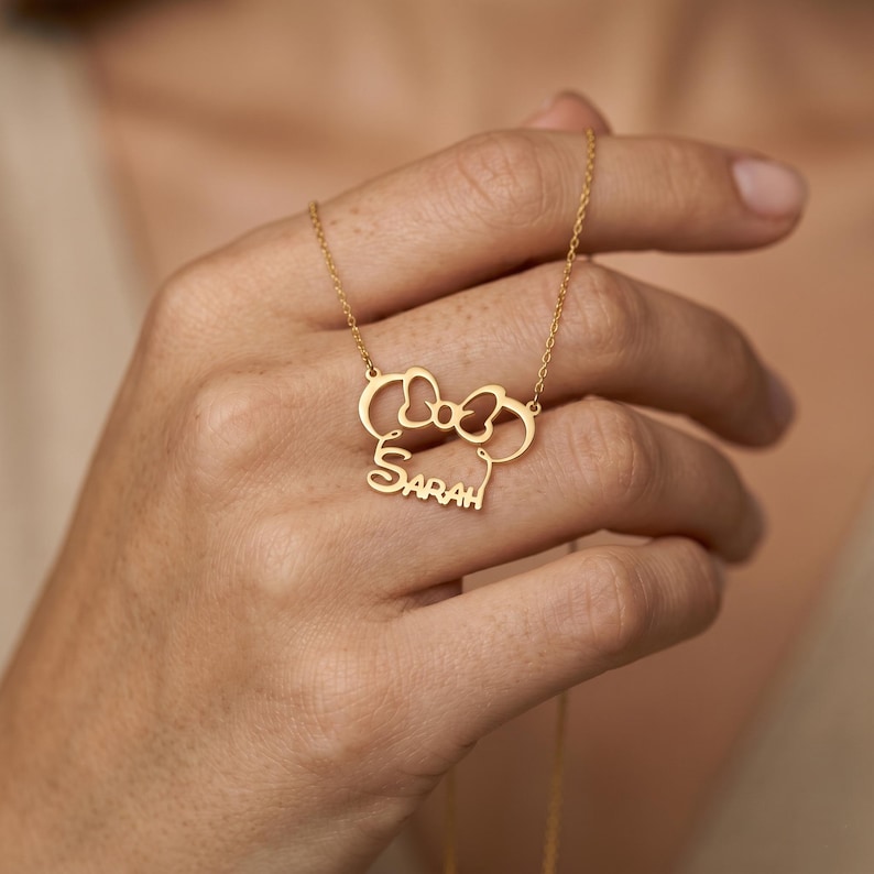 May include: A gold-colored necklace featuring a bow and mouse ears design with the name "Sarah" in a script font. The pendant hangs from a delicate chain. The necklace is held by a person.
