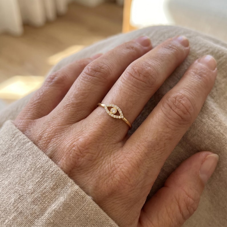 May include: A gold ring featuring an eye-shaped design set with small clear stones and a single central stone. The band is thin and polished, encircling the finger with a delicate, symmetrical aesthetic that highlights the detailed metalwork.