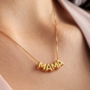 Könnte beinhalten: Eine goldfarbene Halskette mit dem Wort "MAMA" in Großbuchstaben. Der Anhänger hängt an einer feinen Kette. Die Halskette wird um den Hals getragen, wobei sich die goldenen Buchstaben von der Haut abheben.