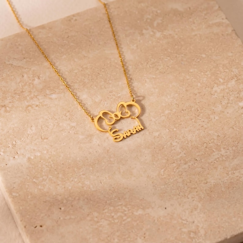 May include: A gold-colored necklace with a bow-shaped pendant and the name "Sarah" in a script font. The necklace is displayed on a light-colored, textured surface. The chain is delicate and the pendant is detailed.