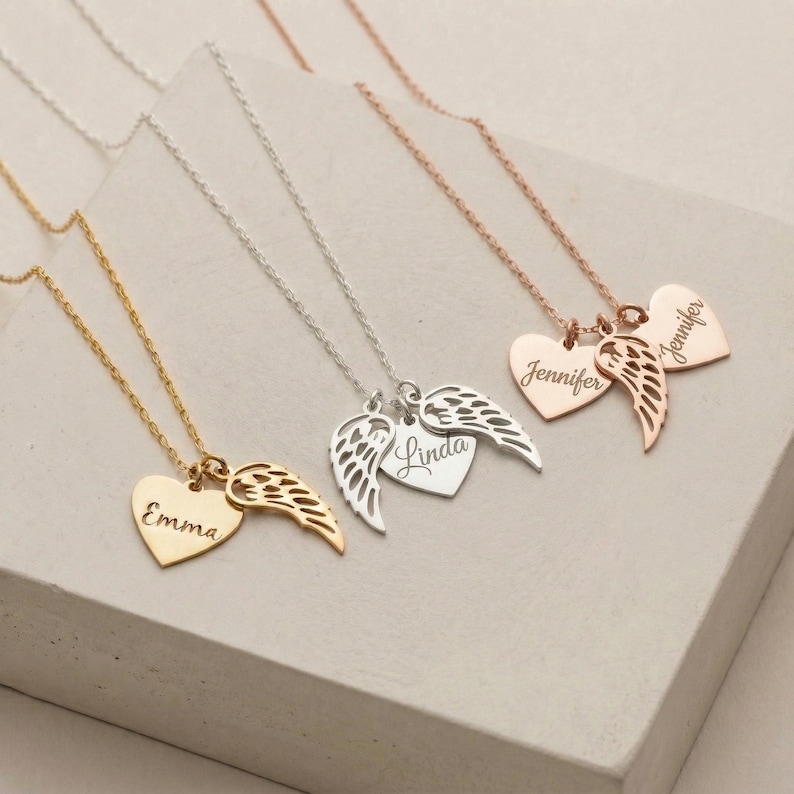 May include: A collection of personalized heart-shaped pendant necklaces in gold, silver, and rose gold. Each necklace features a heart pendant with a name engraved and a wing charm. The names "Emma", "Linda", and "Jennifer" are visible.