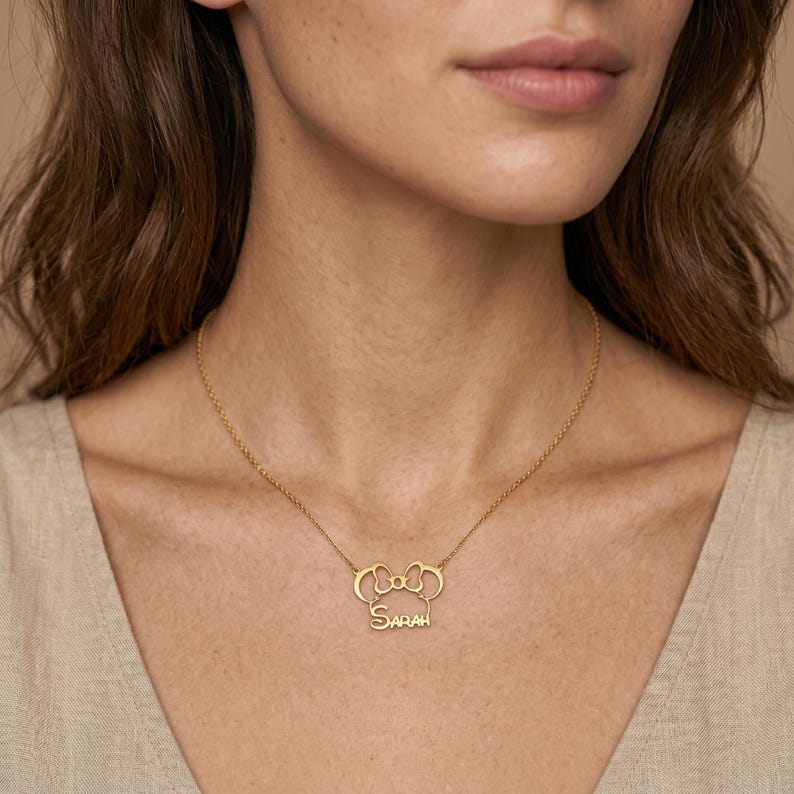 May include: A gold-colored necklace with a pendant shaped like a bow and the name "Sarah" in a script font. The necklace is worn around the neck and is a delicate piece of jewelry.