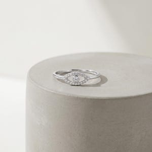 May include: A silver-toned ring featuring an eye-shaped centerpiece. The design includes a central round stone surrounded by a halo of small, clear pav&eacute;-set stones, forming an almond shape that tapers to a thin, smooth, polished metal band.