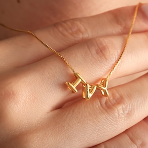May include: A gold-colored necklace with the letters "IVY" as a pendant. The necklace is delicate, with a thin chain and the letters are in a stylized font. The necklace is being held by a person.