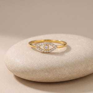 May include: A gold ring featuring an eye-shaped centerpiece. The eye motif is outlined with small, clear, round-cut stones and contains a single, larger, clear, round-cut stone at its center, creating a detailed and polished jewelry piece.