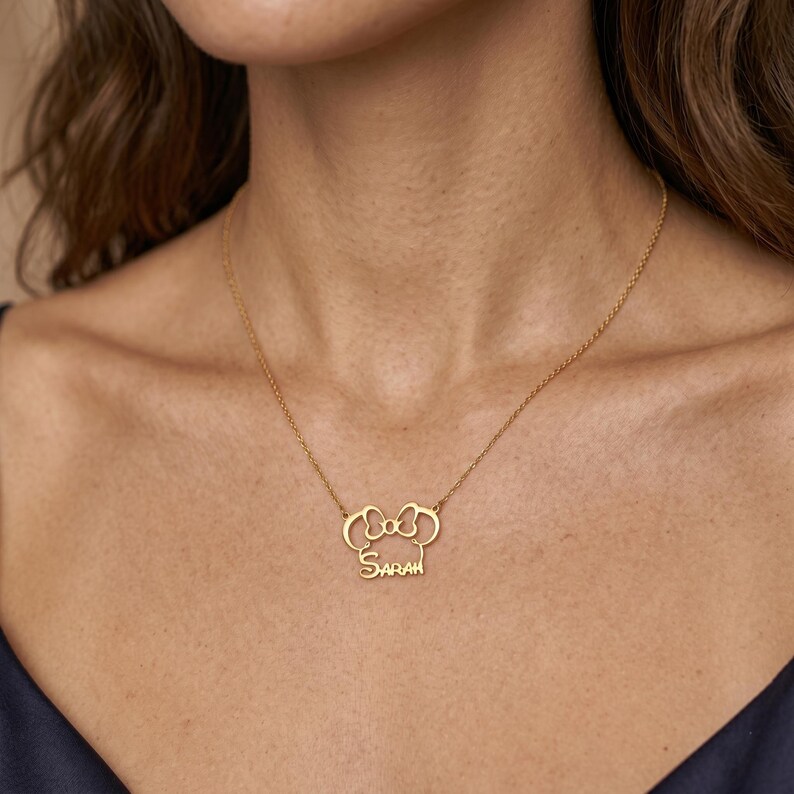 May include: A gold-colored necklace with a bow-shaped pendant and the name "Sarah" in a cursive font. The necklace is worn around a person's neck, with the pendant resting on the chest. The chain is delicate and the pendant is detailed.