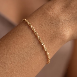 May include: A thin, gold-coloured rope chain bracelet with a twisted, spiral link design. The metal has a polished, reflective finish that catches the light, creating a continuous, textured appearance along the length of the delicate jewellery piece.