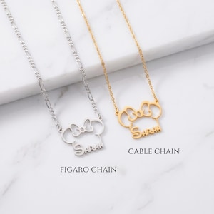 May include: Two necklaces with pendant designs. One is silver with a Figaro chain, the other is gold with a cable chain. Both pendants are shaped like a mouse head with a bow and the name "Sarah" in script. The text "FIGARO CHAIN" and "CABLE CHAIN" are below the necklaces.