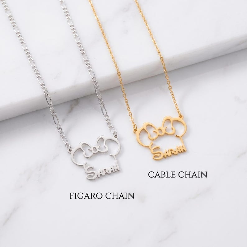 May include: Two necklaces with pendant designs. One is silver with a Figaro chain, the other is gold with a cable chain. Both pendants are shaped like a mouse head with a bow and the name "Sarah" in script. The text "FIGARO CHAIN" and "CABLE CHAIN" are below the necklaces.