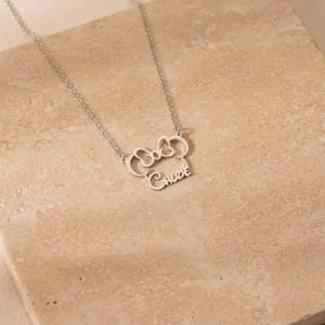 May include: A silver-colored necklace with a bow-shaped pendant and the name "Chloe" in cursive script. The necklace is displayed on a light beige surface, showcasing the delicate design and craftsmanship of the jewelry.
