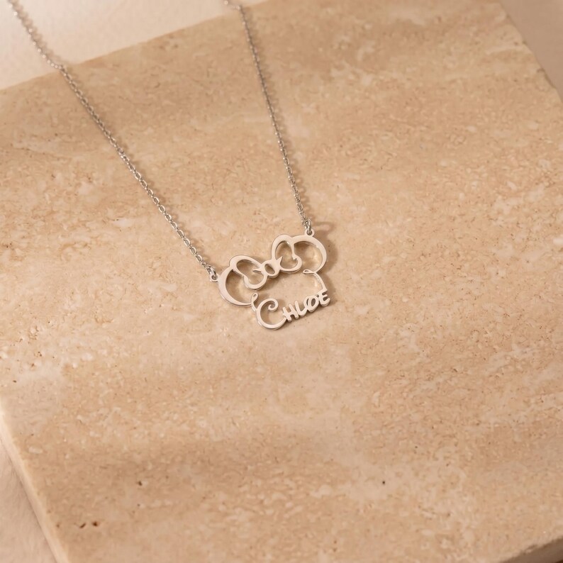 May include: A silver-colored necklace with a bow-shaped pendant and the name "Chloe" in cursive script. The necklace is displayed on a light beige surface, showcasing the delicate design and craftsmanship of the jewelry.