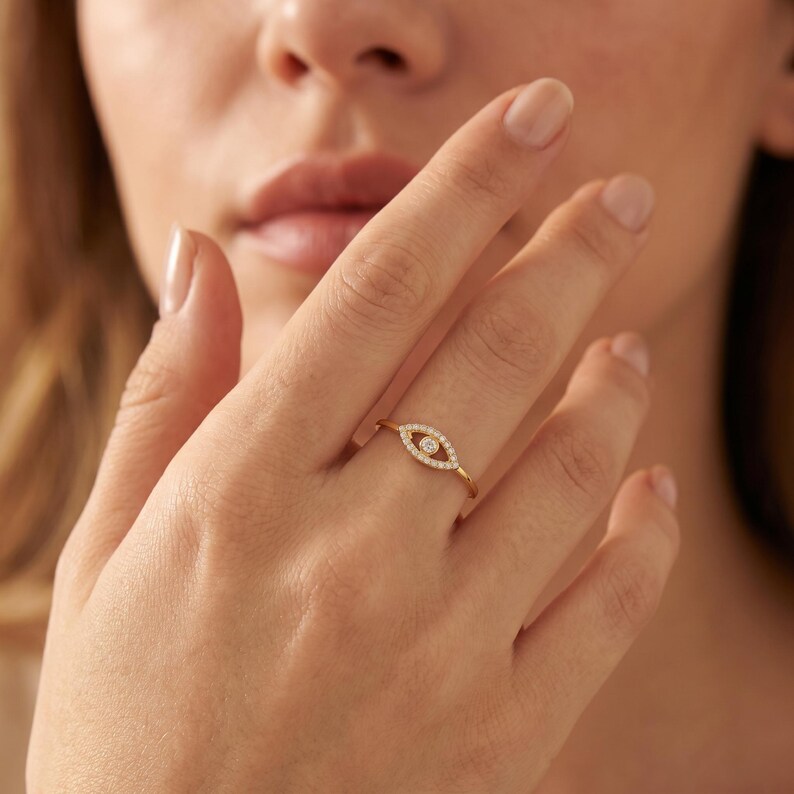 May include: A gold ring featuring an eye-shaped centerpiece. The design includes a central round stone surrounded by a halo of small, clear pav&eacute;-set stones, forming an almond shape. The band is thin, polished gold, creating a delicate appearance.