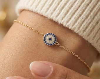 May include: Gold-toned chain bracelet featuring a circular charm with a blue and white evil eye design. The bracelet is worn on a wrist, with a cream-colored sweater in the background. The charm is detailed with small blue and white stones.