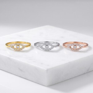 May include: Three rings featuring an eye-shaped centerpiece adorned with small clear stones and a central round stone. The rings are crafted in yellow gold, silver, and rose gold tones, each showcasing a delicate band and a symmetrical design.