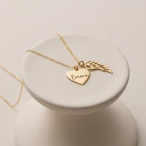 May include: A gold-colored necklace with a heart pendant engraved with the name "Emma" and a wing charm. The necklace is displayed on a white, round, tiered pedestal. The chain and charms are shiny.
