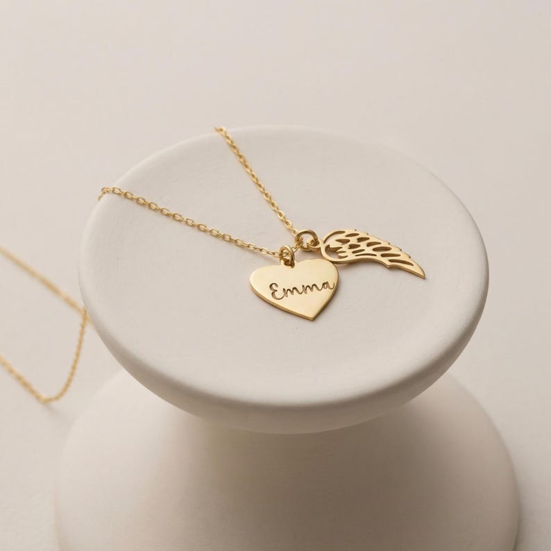 May include: A gold-colored necklace with a heart pendant engraved with the name "Emma" and a wing charm. The necklace is displayed on a white, round, tiered pedestal. The chain and charms are shiny.